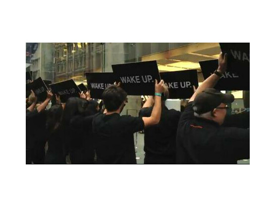 Schwarz gekleidete Menschen halten vor dem Apple Store schwarze Schilder hoch, auf denen schlicht "Wake up" steht.